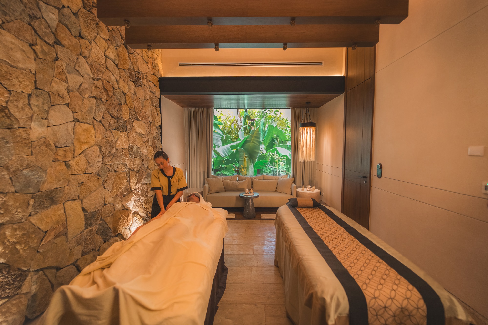 Spa therapist massages a client; stone wall, lush garden view. Hacienda Xcanatun, Angsana Heritage Collection.